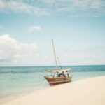 Traditional wooden dhow boat anchored on a sandy beach with turquoise water and footprints in the sand, under a partly cloudy sky in Zanzibar, Tanzania.