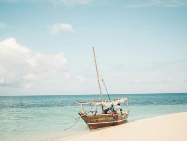 Traditional wooden dhow boat anchored on a sandy beach with turquoise water and footprints in the sand, under a partly cloudy sky in Zanzibar, Tanzania.